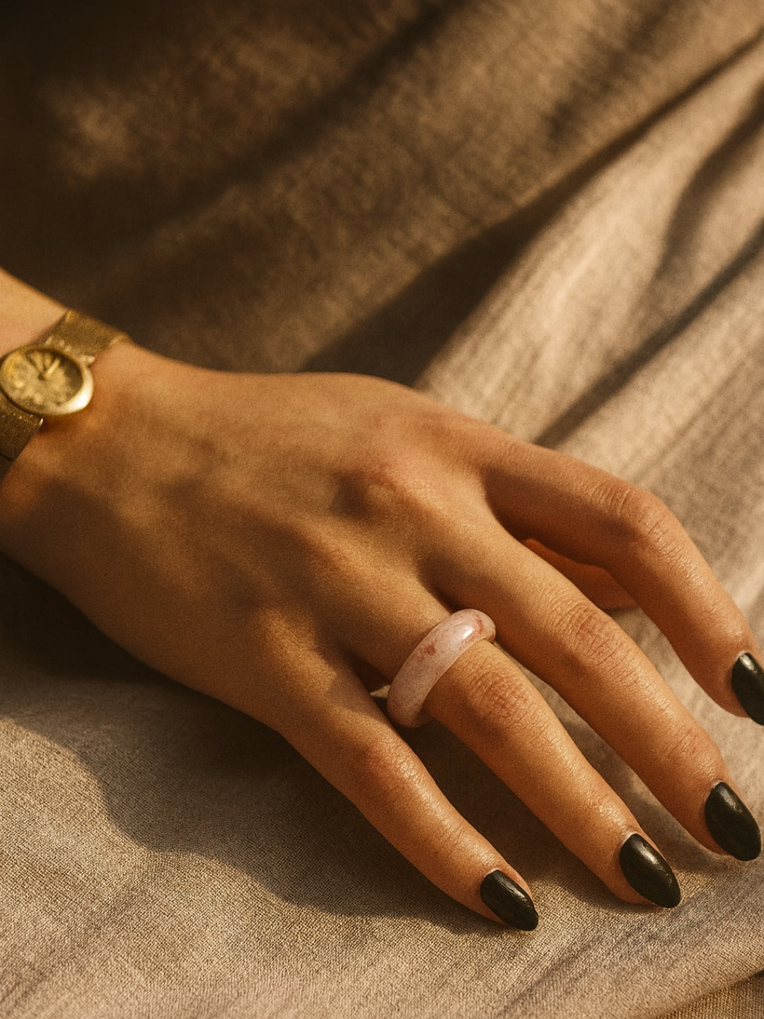 Hand wearing a gold watch and pink ring on a textured surface