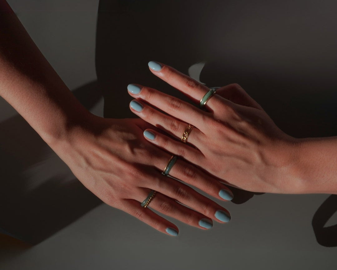 Close-up of two hands wearing multiple jade and gold rings, with soft shadows and muted lighting – Aulyn handcrafted jewelry collection.