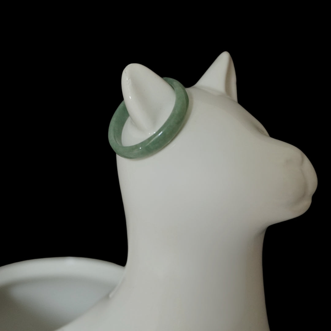 Light green Jade ring on white ceramic cat jewelry holder, minimalist product display on black background.