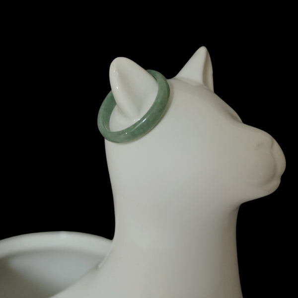 Light green Jade ring on white ceramic cat jewelry holder, minimalist product display on black background.