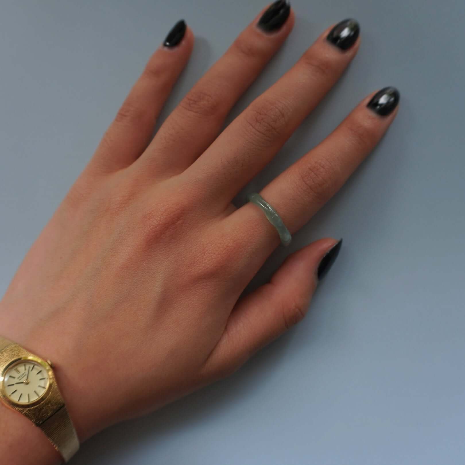 Hand wearing a minimalist green jade ring paired with a vintage gold wristwatch, displayed against a neutral blue-gray background for jewelry styling context.