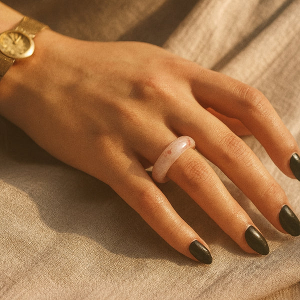 Hand wearing a minimalist pink agate ring on the ring finger, paired with a vintage gold wristwatch, displayed against a neutral blue-gray background for jewelry styling context.