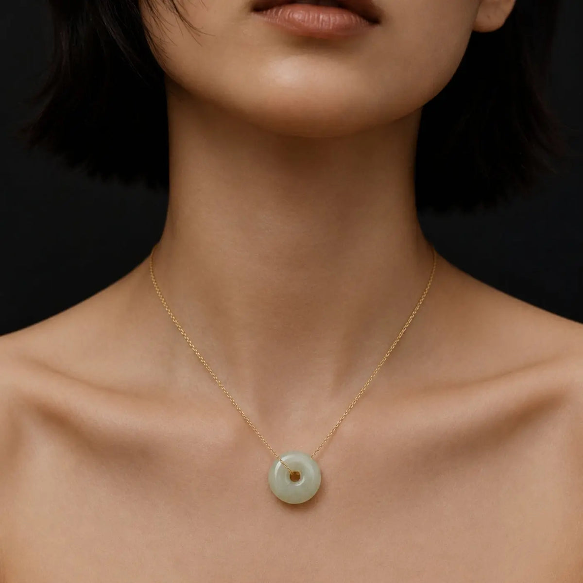 Close-up portrait of a woman wearing a delicate gold chain necklace with a pale green jade donut pendant against a dark studio background.