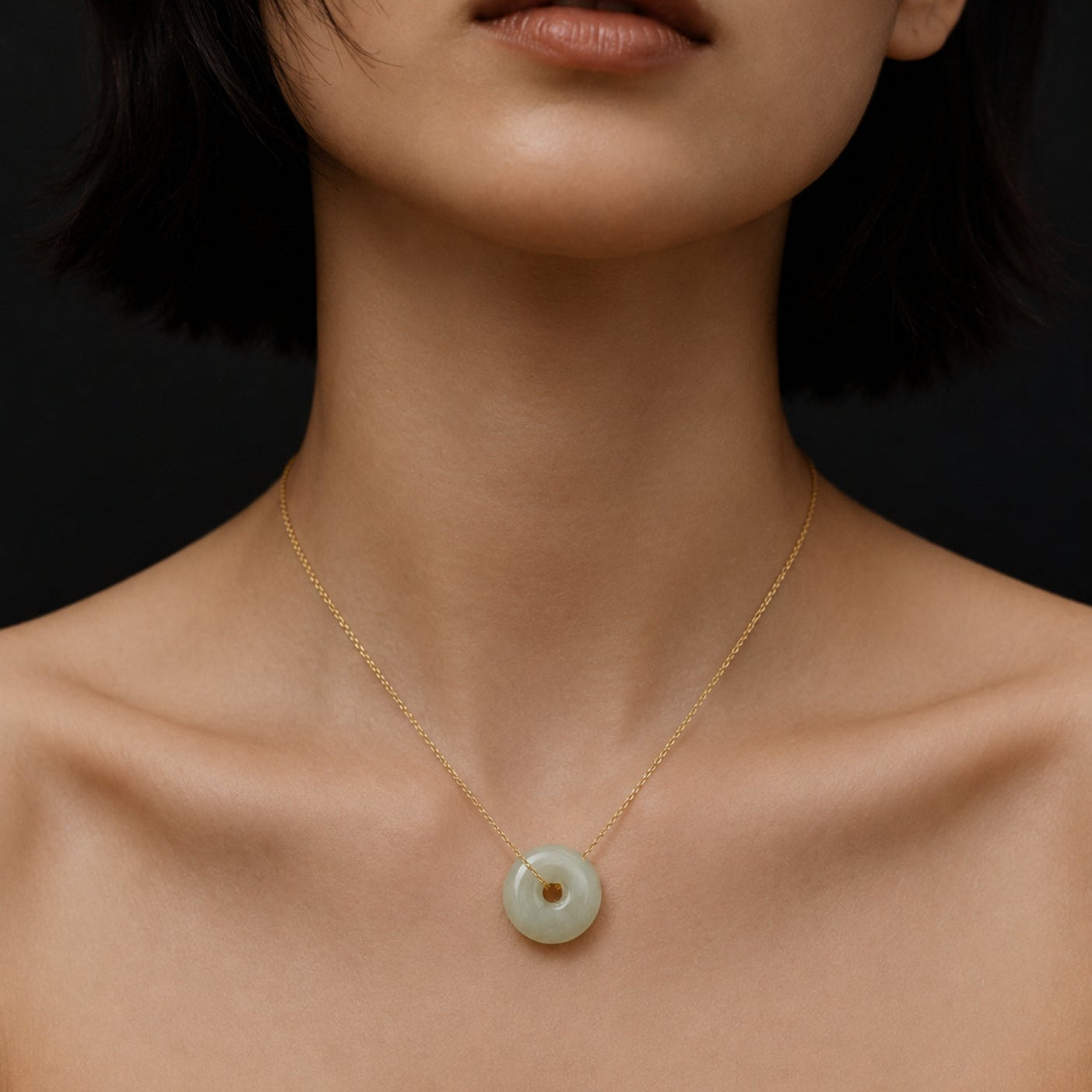 Close-up portrait of a woman wearing a delicate gold chain necklace with a pale green jade donut pendant against a dark studio background.