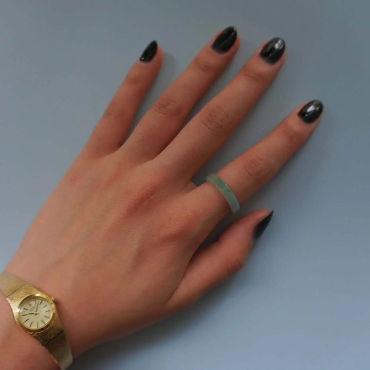 Hand wearing a minimalist green jade ring on the ring finger, paired with a vintage gold wristwatch, displayed against a neutral blue-gray background for jewelry styling context.