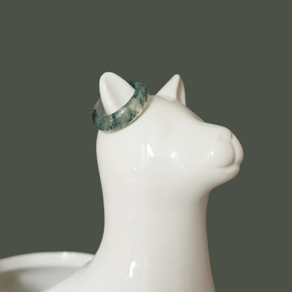 Light green Moss Agate ring on white ceramic cat jewelry holder, minimalist product display on green background.