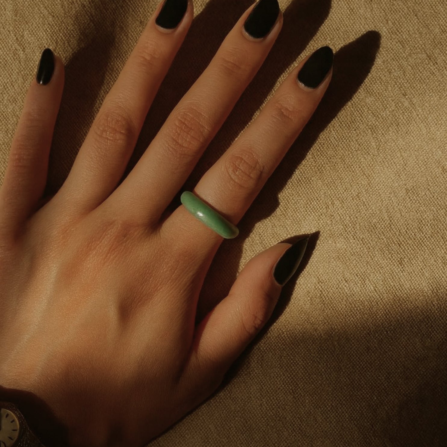 Hand wearing a minimalist green jade ring, paired with a vintage gold wristwatch, displayed against a neutral blue-gray background for jewelry styling context.